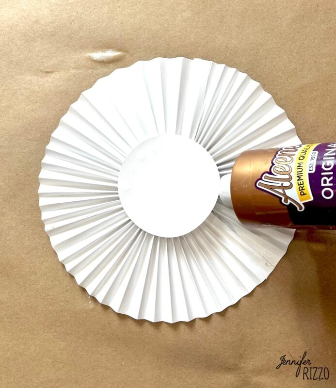 A bottle of Aleene's Tacky Glue is positioned above the center of a folded white paper rosette on a brown surface.