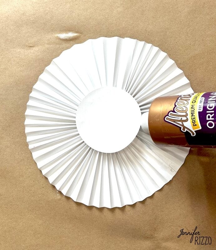 A bottle of Aleene's Tacky Glue is positioned above the center of a folded white paper rosette on a brown surface.