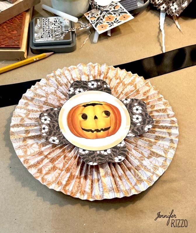 A paper rosette decoration features a smiling jack-o’-lantern in the center, surrounded by patterned paper petals, on a craft table with various supplies.