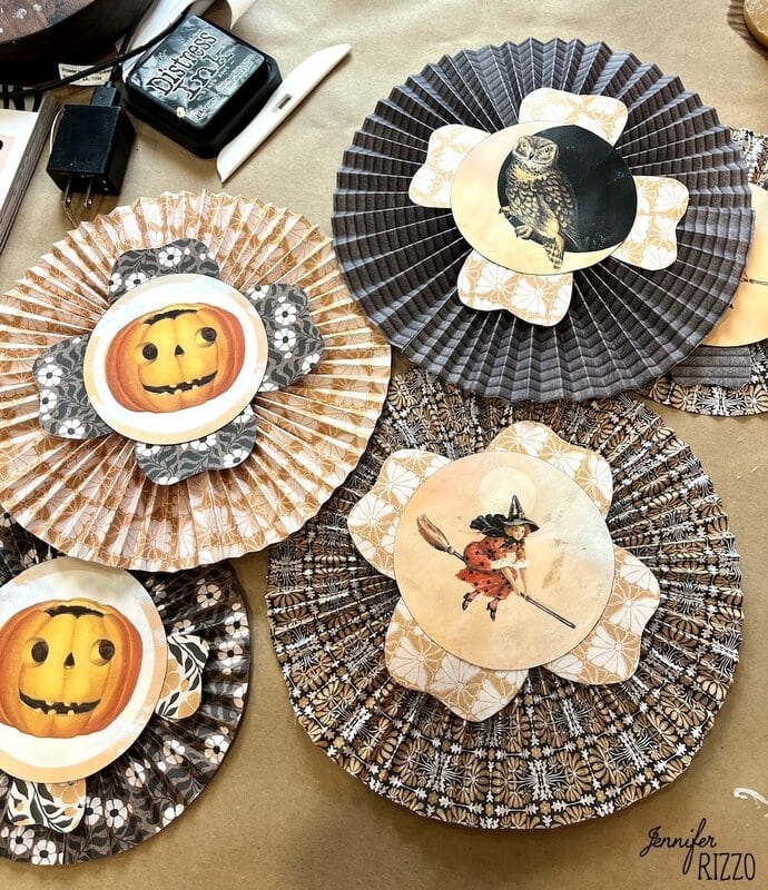 Four decorative paper rosettes with vintage Halloween images, including a pumpkin, owl, and a witch, displayed on a craft table with ink and other supplies.