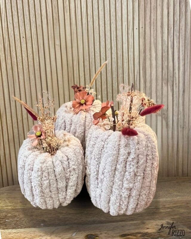 Three soft, knitted pumpkins with dried flowers and decorative stems arranged on a wooden surface against a light wood panel background.
