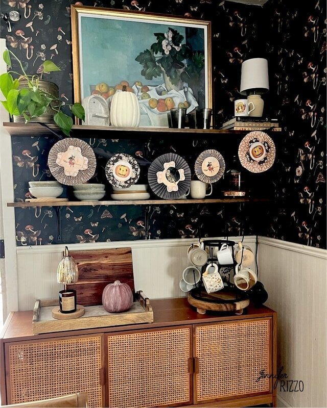 A kitchen corner with dark floral wallpaper, wooden shelves displaying dishware and decorative plates, and a sideboard with pumpkins and a tray beneath.
