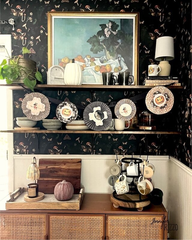 A kitchen corner with shelves holding dishes, mugs, decor, a framed fruit painting, and Halloween-themed paper rosettes hanging on the middle shelf. A purple pumpkin sits on the counter.