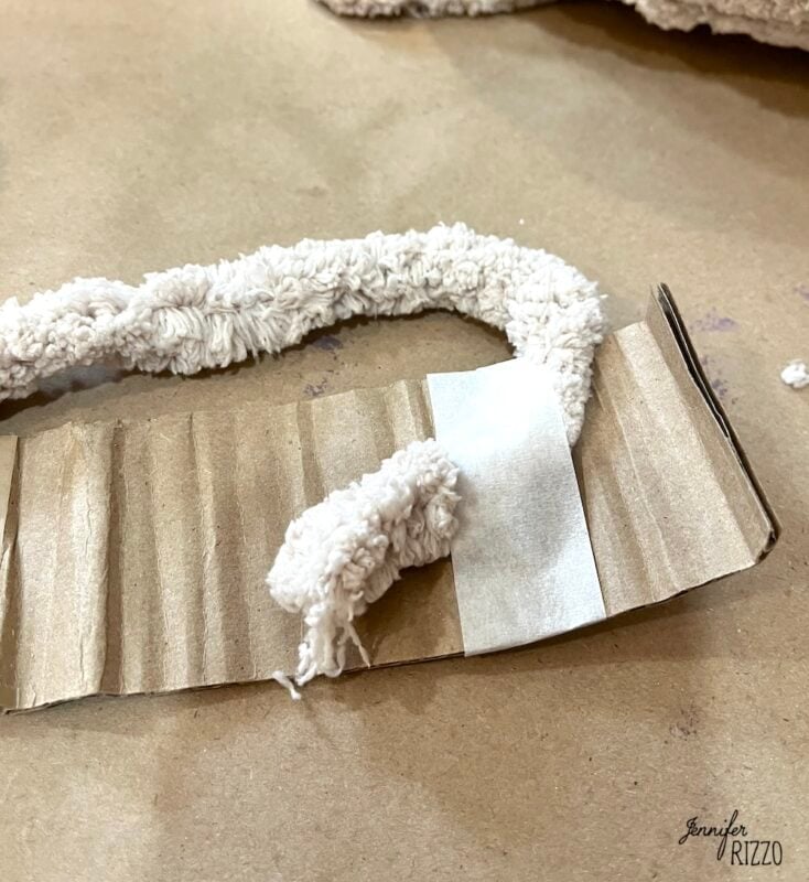 A piece of textured cream-colored yarn is taped to a strip of corrugated cardboard on a brown surface.