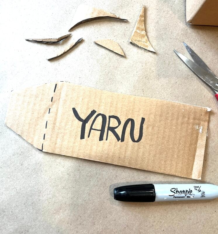 A piece of cardboard labeled "YARN" with a Sharpie marker, scissors, and cardboard scraps on a table.
