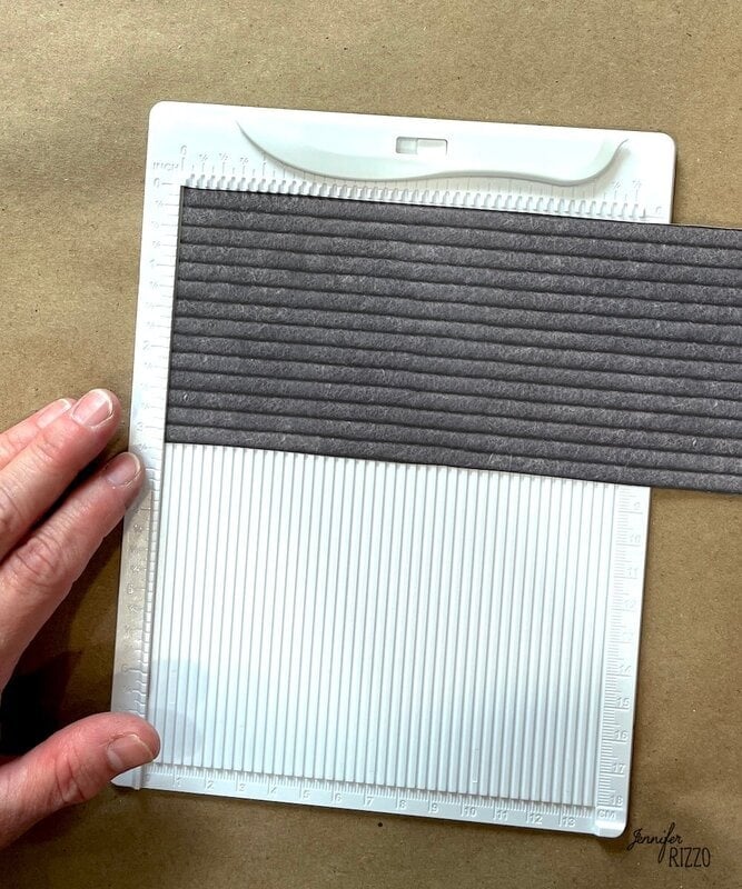A hand holds a piece of dark cardstock horizontally on a white scoring board with evenly spaced grooves, against a brown background.
