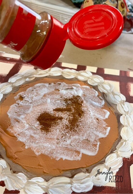 A hand sprinkles cinnamon from a red-topped container onto a round, creamy surface with a decorative border, resembling the cozy charm of Pumpkin Pie Wall Art or a festive dessert.