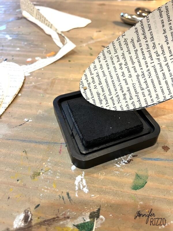 A paper heart cutout with printed text is being pressed onto a black ink pad on a wooden table with crafting materials.