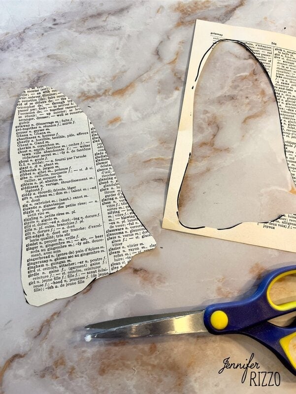 A pair of scissors, a cut-out dictionary page in the shape of a bell, and a matching negative space piece rest on a marble surface.