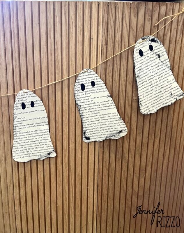Three ghost shapes cut from printed book pages are strung on twine and hung as a garland against a wooden background.