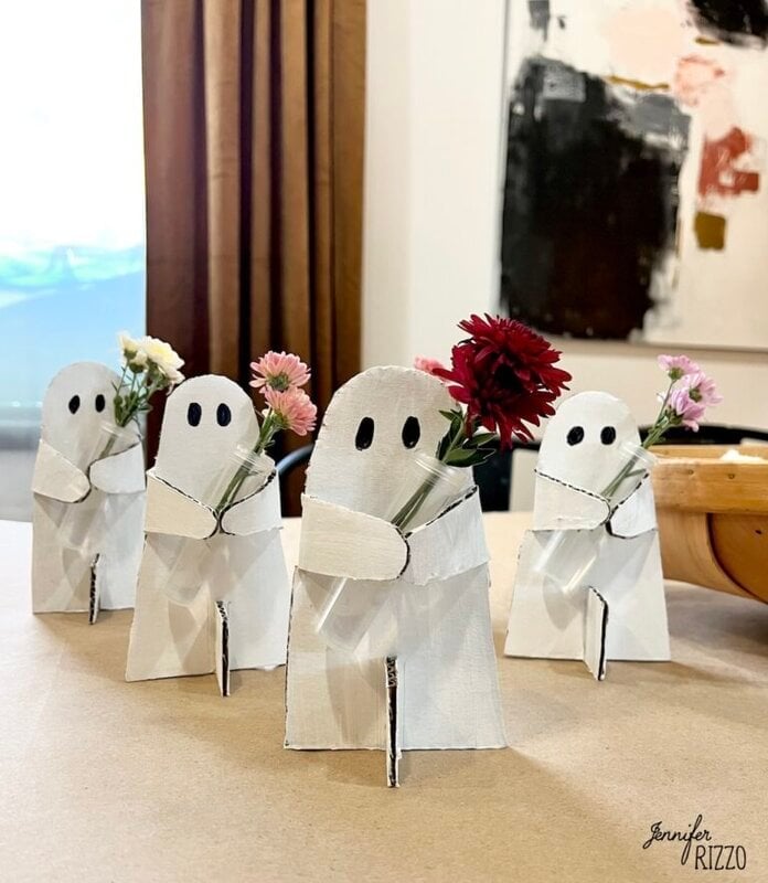Four small ghost-shaped paper crafts each hold a flower, arranged on a table with a blurred background of a window, curtains, and wall art.