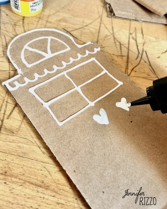 A cardboard cutout shaped like a house being decorated with white puff paint to create windows and hearts.