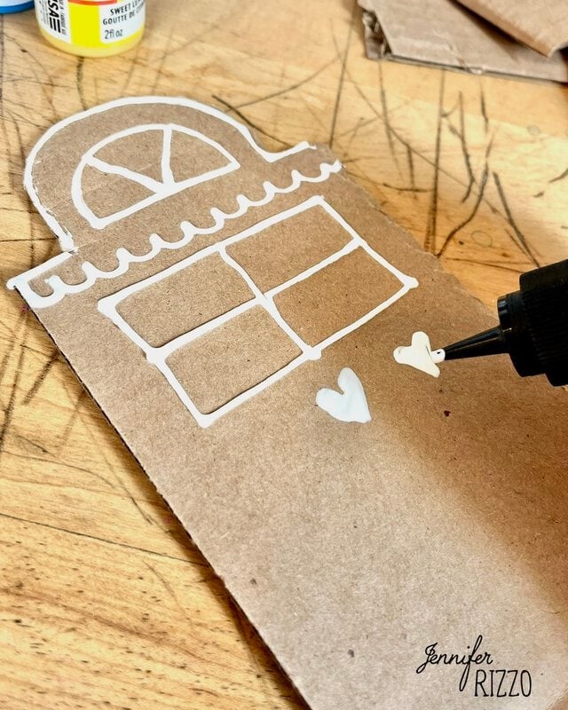A cardboard cutout shaped like a house being decorated with white puff paint to create windows and hearts.
