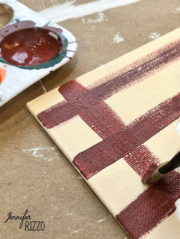 A paintbrush applies red paint in a plaid pattern on a canvas, creating Pumpkin Pie Wall Art; a palette with paint rests nearby on a brown work surface.