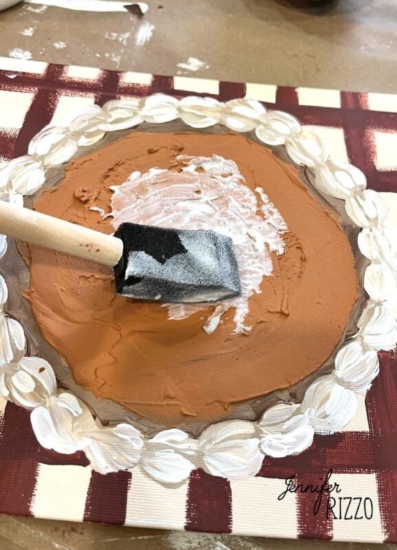 A foam brush applies white paint to the center of a brown painted circle, resembling Pumpkin Pie Wall Art, with a white floral border on a red and white checkered canvas.