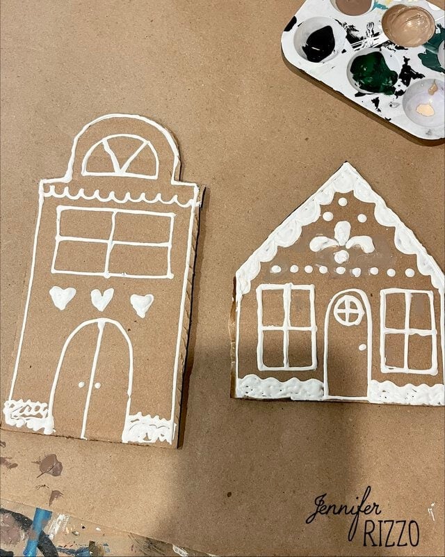 Two cardboard gingerbread house cutouts decorated with white puffy paint, next to a paint palette.