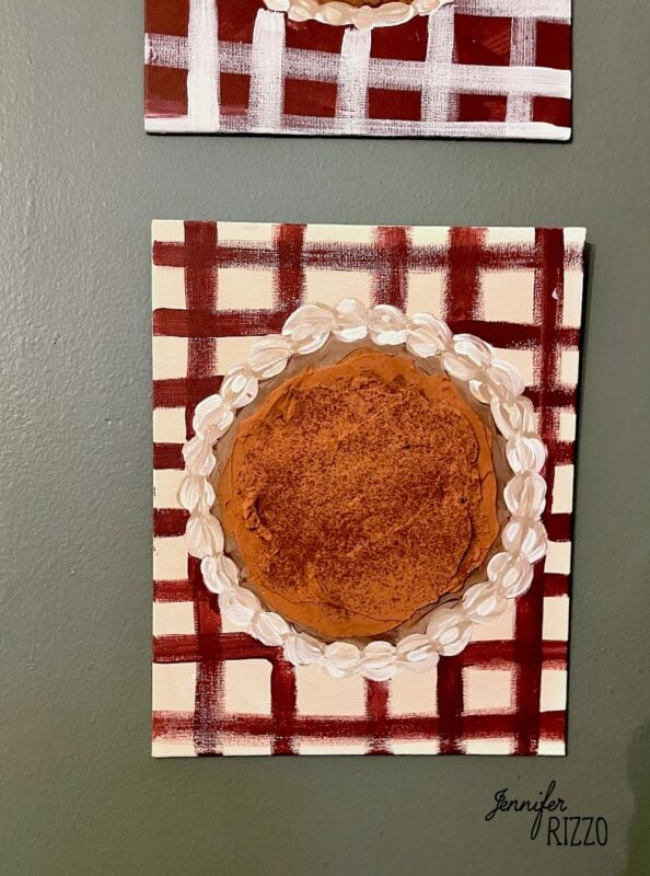 A painting of a pumpkin pie with a decorative whipped cream border, set on a red and white plaid background. This charming Pumpkin Pie Wall Art is signed by artist Jennifer Rizzo in the bottom right corner.