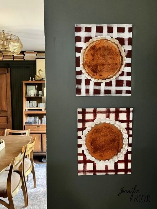 Two square Pumpkin Pie Wall Art pieces with lattice backgrounds hang vertically on a dark wall in a dining area featuring wood furniture and shelves in the background.