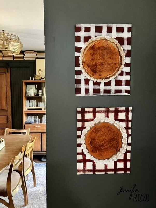 Two square Pumpkin Pie Wall Art pieces with lattice backgrounds hang vertically on a dark wall in a dining area featuring wood furniture and shelves in the background.