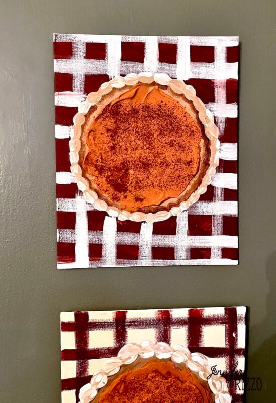 A painting of a pumpkin pie with a decorative crust, set on a red and white checkered tablecloth background, hangs on a dark wall—perfect Pumpkin Pie Wall Art for your kitchen or dining area.