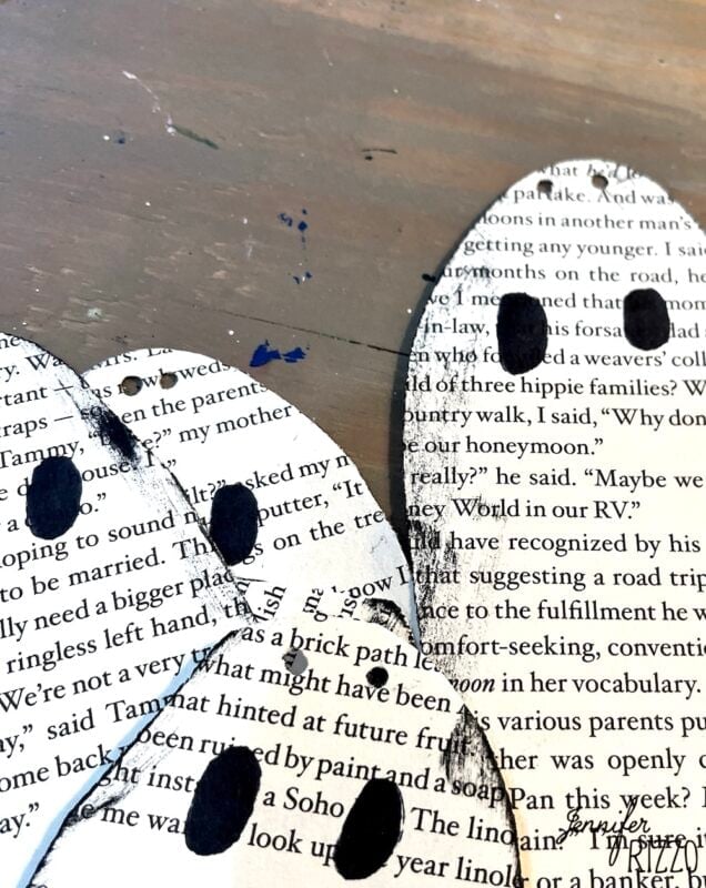 Three paper cutouts shaped like ghosts feature text from book pages and have simple black eyes, placed on a wooden surface.