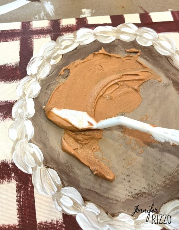 A palette knife mixes brown and tan paint on a painted plate with a white leaf border, atop a red and cream checkered surface. This charming Pumpkin Pie Wall Art is signed "Jennifer Rizzo.