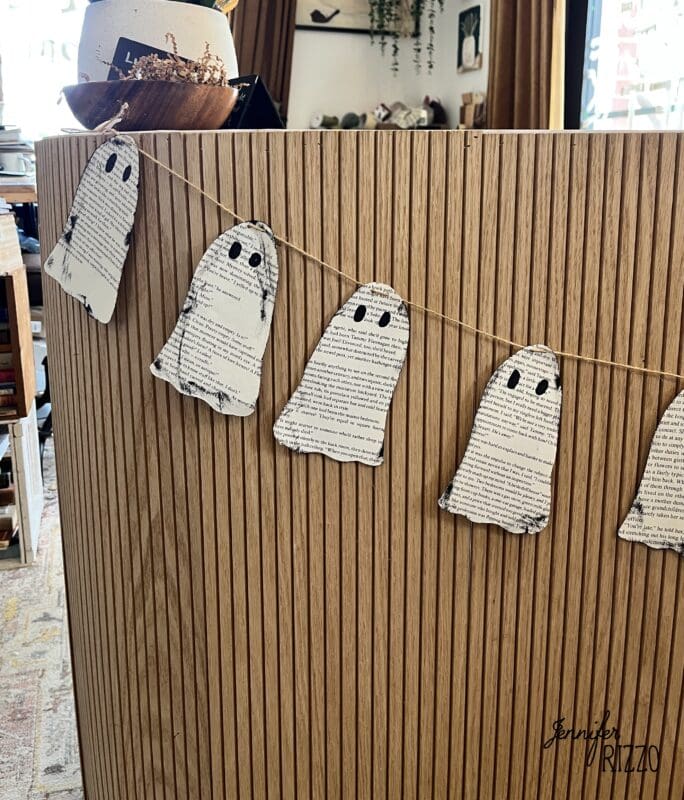 String of ghost-shaped cutouts made from printed paper, hanging as a garland on a wooden slatted surface in a decorated indoor space.