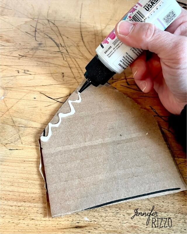 A hand applying a line of white glue along the edge of a cardboard cutout.