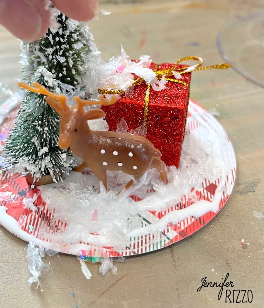 A small plastic deer, a glittery red gift box, and a snowy tree are arranged on a decorated plate with artificial snow.