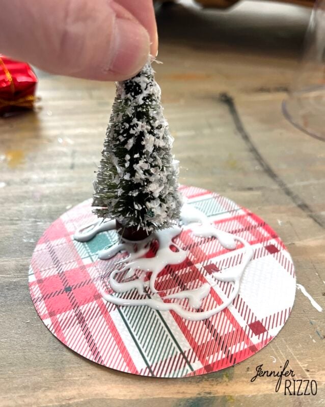 A hand places a small, snow-covered artificial tree onto a plaid-patterned circle with visible white glue underneath.