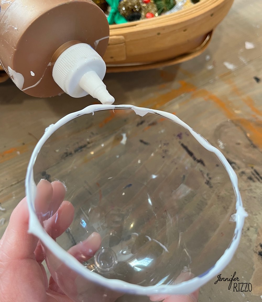 A hand holds a clear plastic cup with white glue applied around the rim; an open glue bottle and craft materials are visible in the background.