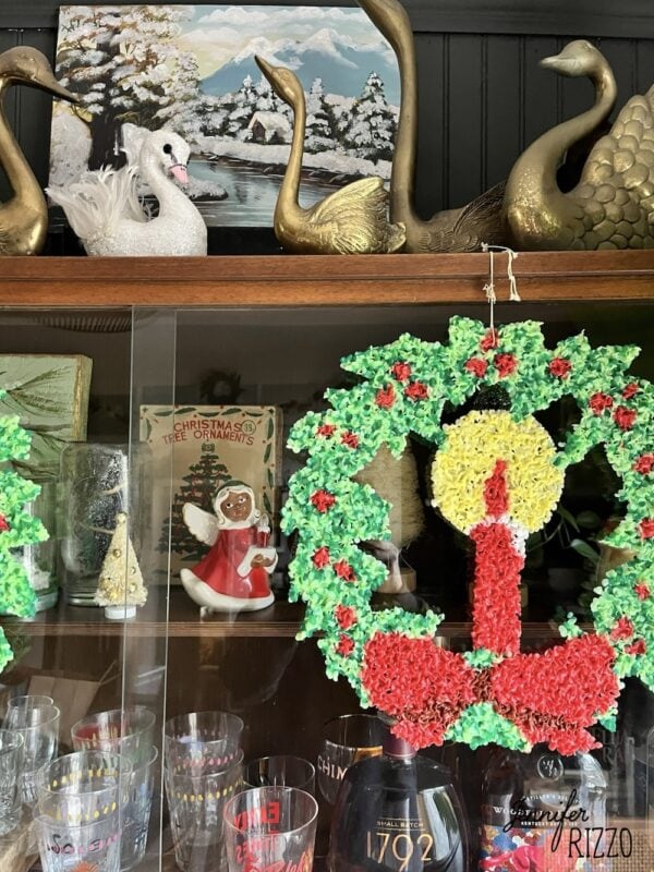 A glass cabinet displays nostalgic and vintage Christmas decorating touches, featuring a wreath with a candle, glassware, a ceramic angel, swan figurines, and a winter landscape painting.