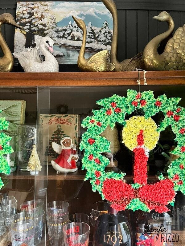 A glass cabinet displays nostalgic and vintage Christmas decorating touches, featuring a wreath with a candle, glassware, a ceramic angel, swan figurines, and a winter landscape painting.