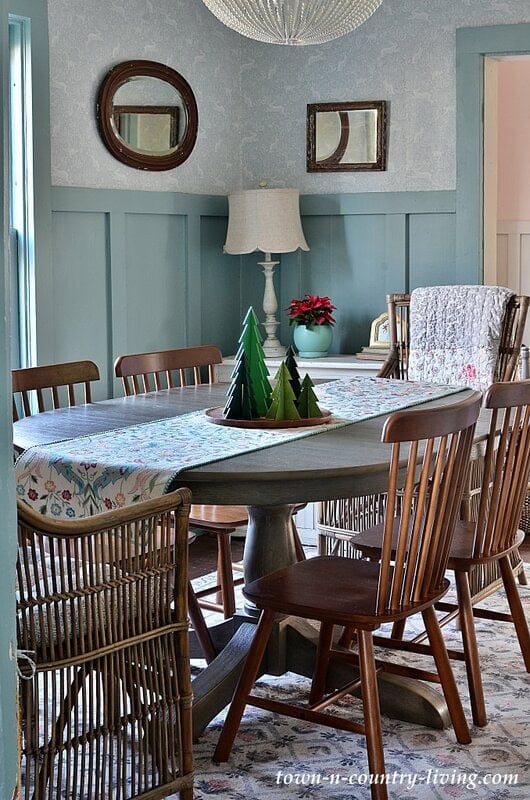 A dining room with a round table, mixed wooden chairs, a floral tablecloth, fabric trees centerpiece, a lamp, a small poinsettia, and decorative mirrors on blue paneled walls.