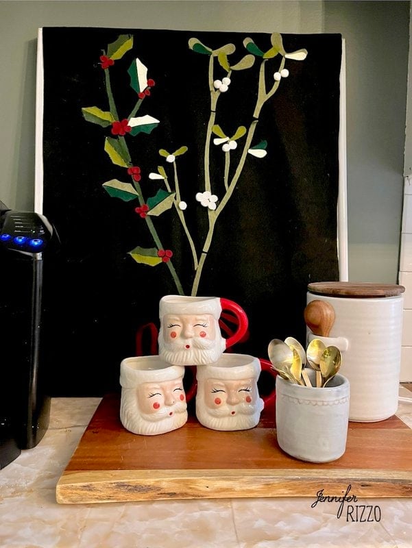 Three Santa-themed mugs, a white ceramic jar with spoons, and a canister are arranged on a wooden tray in front of festive botanical artwork—perfect touches for nostalgic and vintage Christmas decorating.