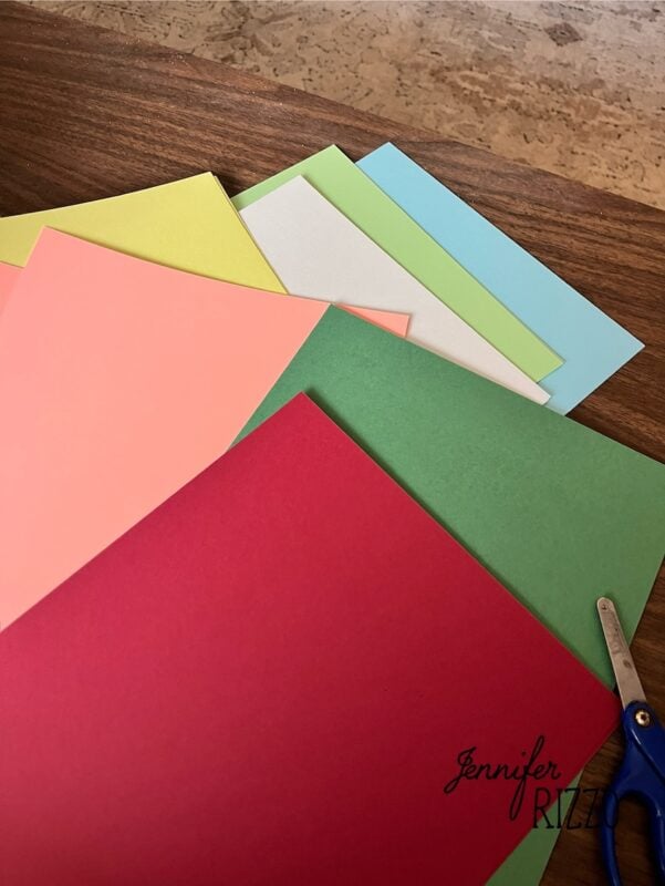 Several sheets of colored paper in red, green, pink, yellow, white, and blue are spread out on a wooden surface next to a pair of scissors.