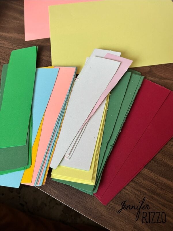 A stack of colorful paper strips and sheets, including green, yellow, blue, pink, gray, and red, arranged on a wooden surface. The name "Jennifer Rizzo" is visible in one corner.