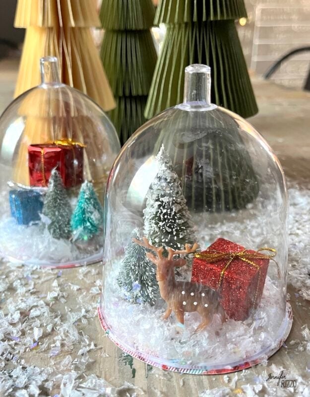 Miniature holiday scene with a plastic dome covering a small deer, trees, artificial snow, and red gift boxes; folded paper trees in the background to make Mini Snow Globe Cloches from Plastic Wine Glasses