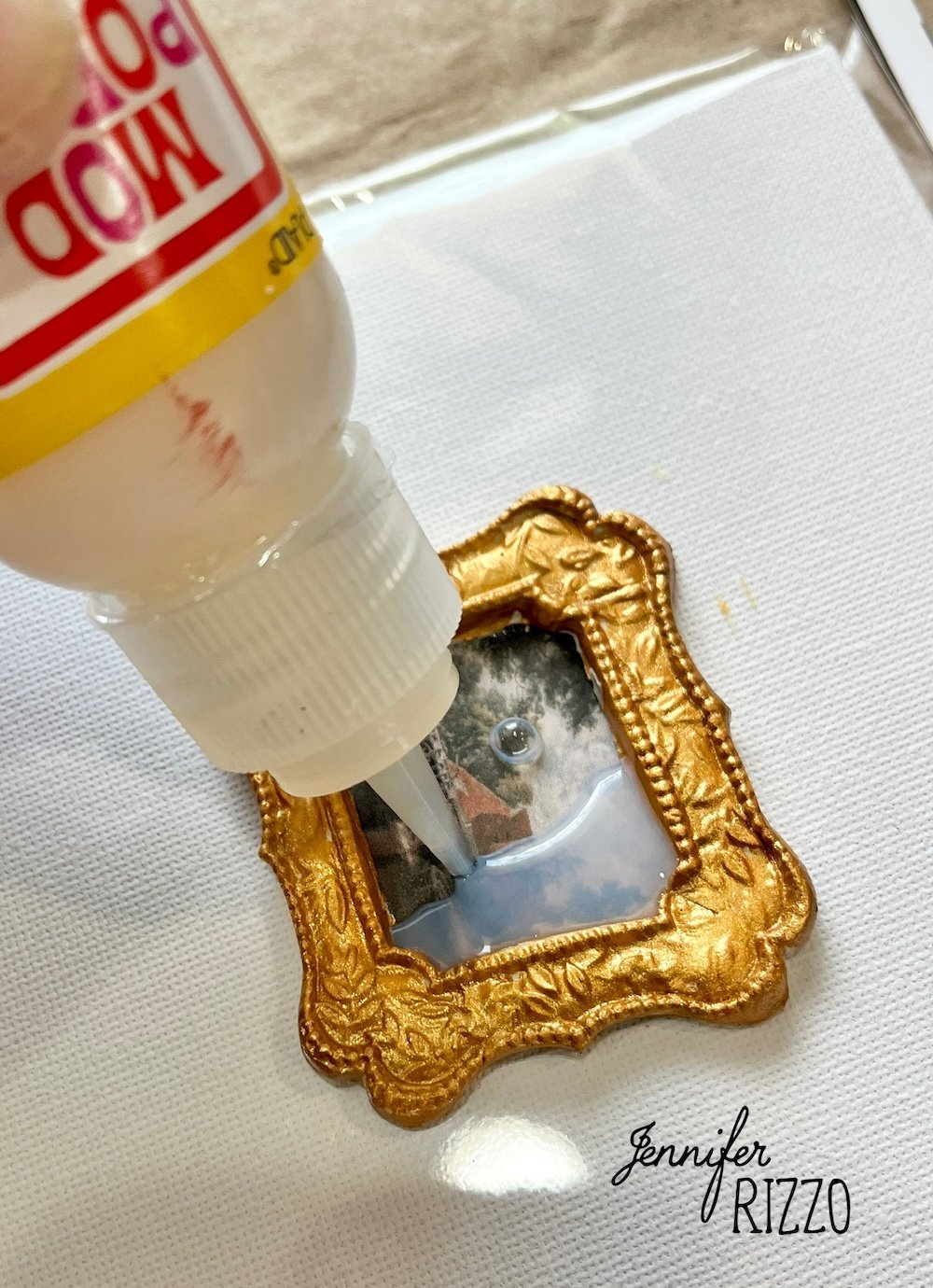 A bottle of Mod Podge is being applied to a small framed picture on a white surface, with "Jennifer Rizzo" written at the bottom right.
