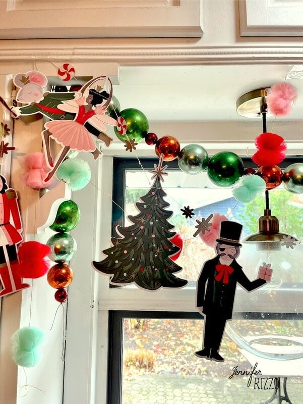 Nostalgic and Vintage Christmas Decorating shines in paper cutouts of a ballerina, Christmas tree, and man in a top hat hanging on a festive garland with green, red, gold ornaments and pastel pom-poms above a window.