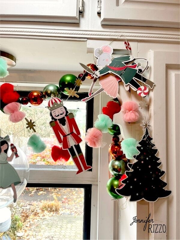 A kitchen cabinet corner decorated with holiday garland, nostalgic and vintage Christmas decorating touches, ornaments of a nutcracker, ballerina mouse, and Christmas tree, finished with red and green pom-poms.
