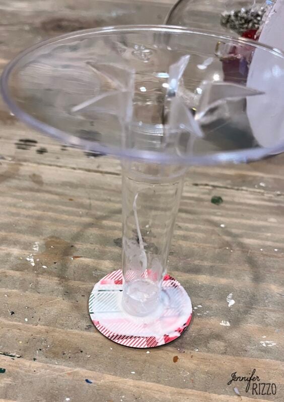 A clear plastic stem and base, with the bottom attached to a round, patterned paper circle, placed on a wooden surface.