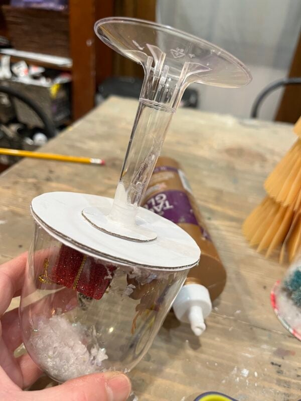 A hand holds an upside-down wine glass with white cardboard glued to the base, containing small holiday decorations and faux snow inside the glass.