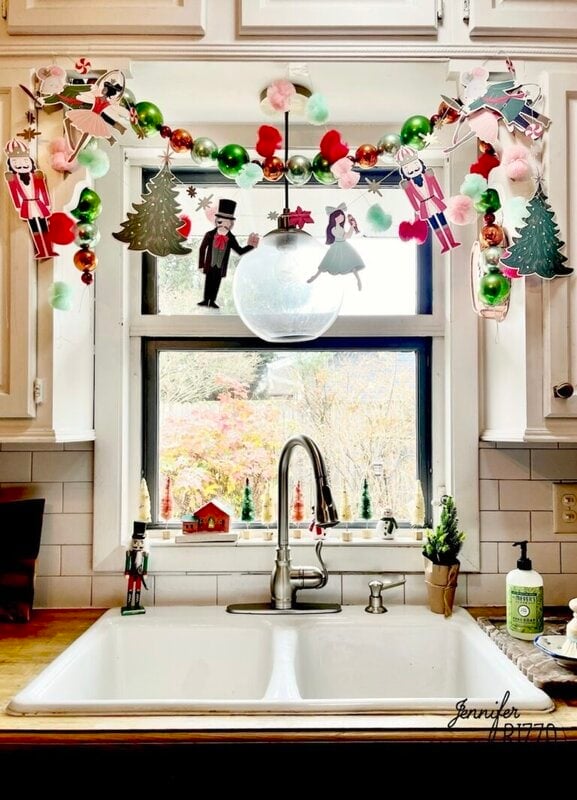 A kitchen sink beneath a window decorated with nostalgic and vintage Christmas garlands, paper figures, green ornaments, and holiday figurines on the windowsill.