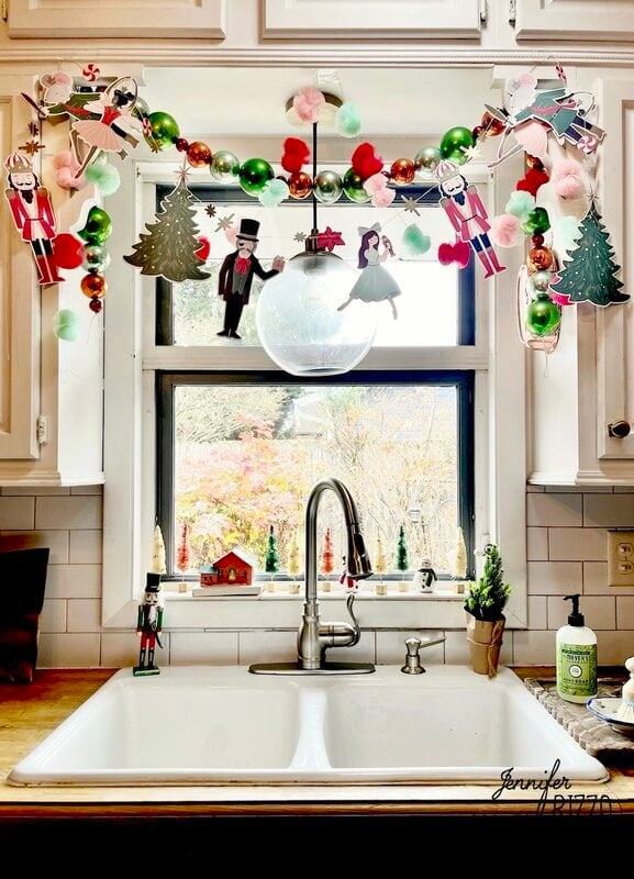 A kitchen sink beneath a window decorated with nostalgic and vintage Christmas garlands, paper figures, green ornaments, and holiday figurines on the windowsill.