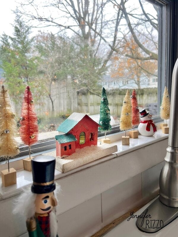 Nostalgic and Vintage Christmas Decorating comes alive in this miniature holiday village display, featuring a red house, bottle brush trees, a snowman, and a nutcracker on a windowsill overlooking wintery trees and a fence outside.