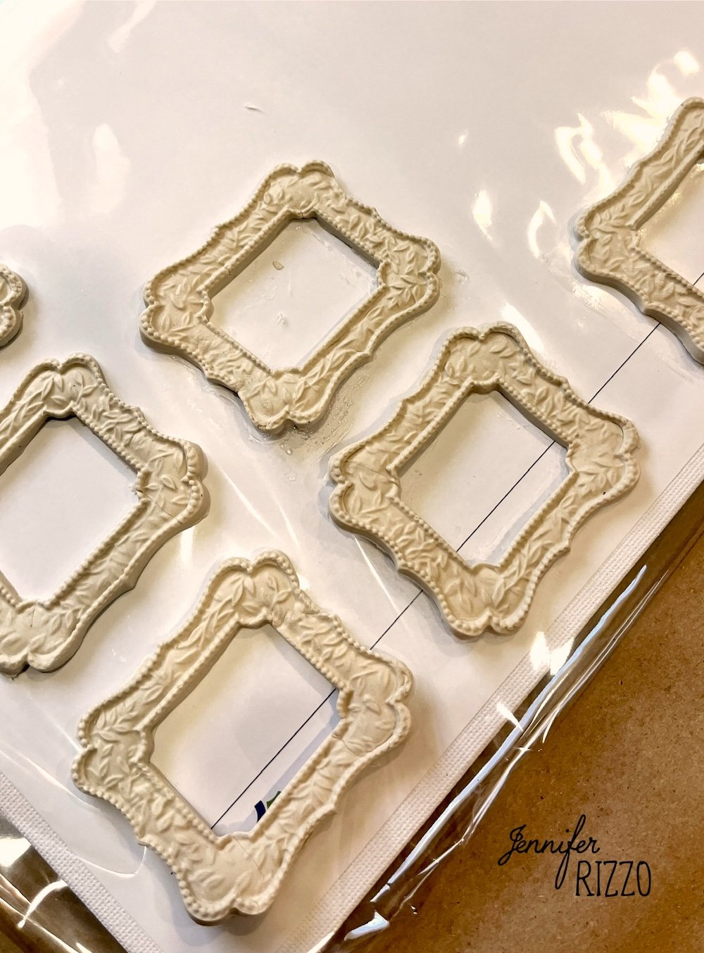 Six small, ornate, cream-colored picture frame embellishments arranged on a clear plastic sheet, with the name "Jennifer Rizzo" visible in the lower right corner.