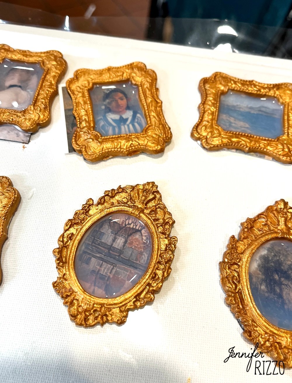 Several ornate miniature gold frames display small classical art prints on a white surface, with the name "Jennifer Rizzo" in the bottom right corner.