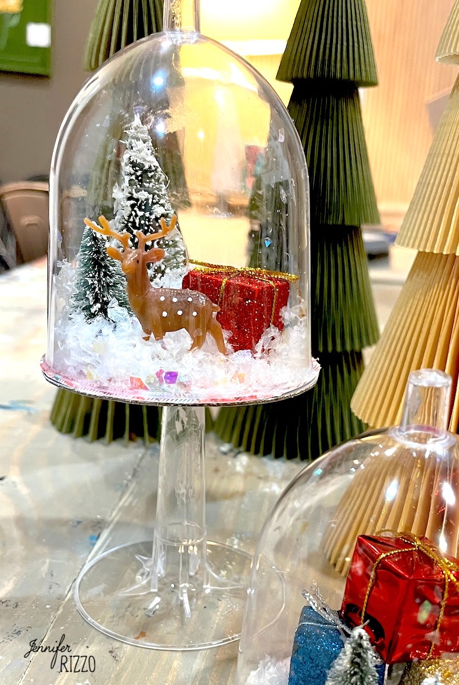 A small deer figurine, gift boxes, and snowy trees are displayed inside an upside-down wine glass, creating a winter scene decoration.
