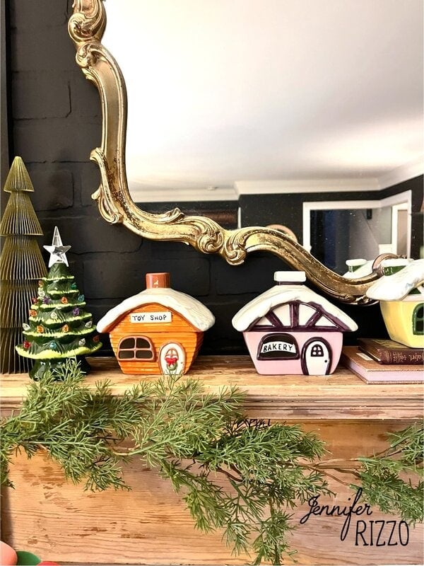 Mini ceramic houses labeled "Toy Shop" and "Bakery" evoke nostalgic and vintage Christmas decorating, displayed on a mantel with greenery, a small Christmas tree, and a decorative mirror in the background.